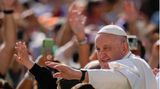 Papst Franziskus winkt in weißem Gewand mit links in eine Menschenmenge