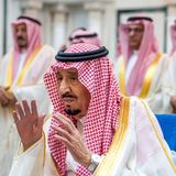 Mit auf Kopfhöhe erhobenen Händen sitzt ein Mann mit rot-weißem Kopftuch und weißem Gewand auf einem blau gepolsterten Stuhl