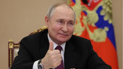 Wladimir Putin sitzt an einem Schreibtisch und hebt lächelnd den Daumen seiner rechten Hand