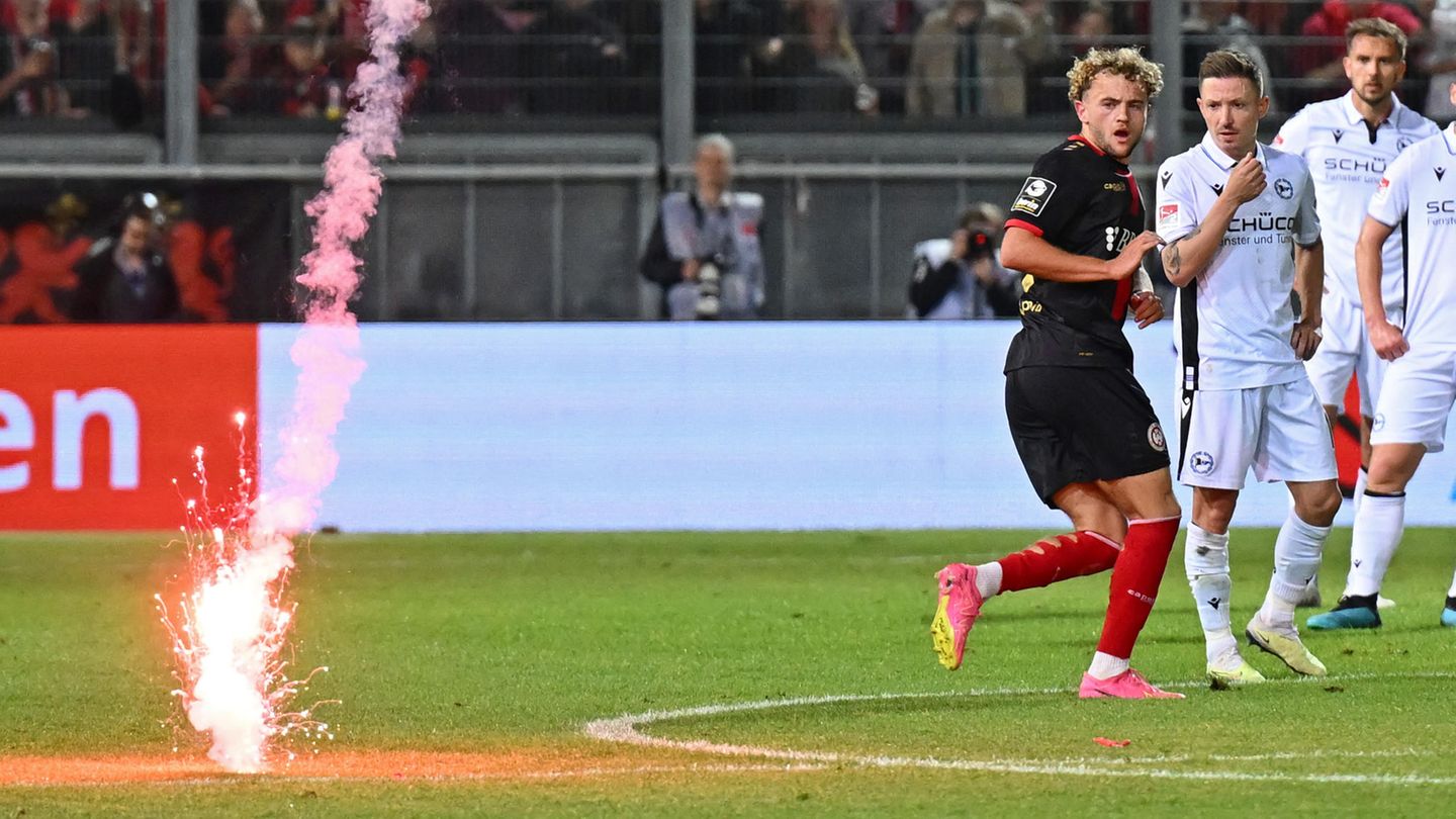 Eine rote Rakete schlägt neben Profi-Fußballern von Wehen Wiesbaden und Arminia Bielefeld auf dem Rasen ein