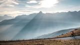 Sanft scheint die Sonne auf die Alpen. Davor liegt ein Weg, auf dem Radfahrer anhält