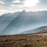 Sanft scheint die Sonne auf die Alpen. Davor liegt ein Weg, auf dem Radfahrer anhält