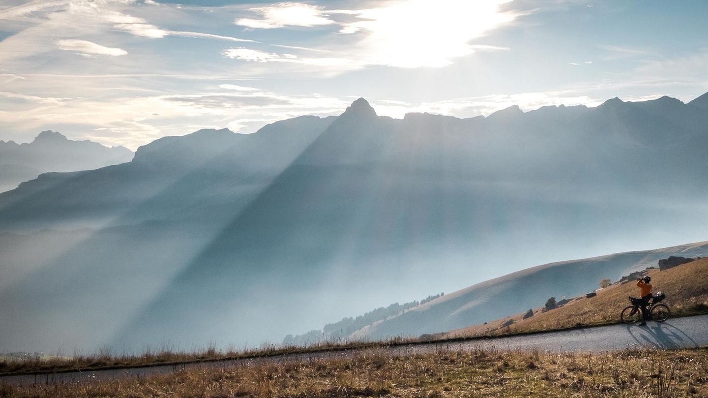 Sanft scheint die Sonne auf die Alpen. Davor liegt ein Weg, auf dem Radfahrer anhält