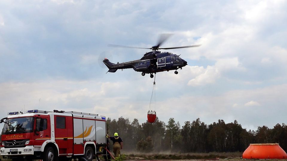Ein Hubschrauber hebt nahe Jüterbog mit einem Wassersack neben einem Feuerwehrwagen ab