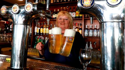 Auf Mallorca steht eine blonde Frau mit drei Bierkrügen in der Hand lächelnd hinter einem Tresen
