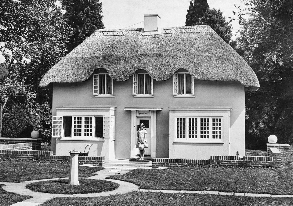 A small house with a thatched roof, in front of which the Queen stands as a young girl