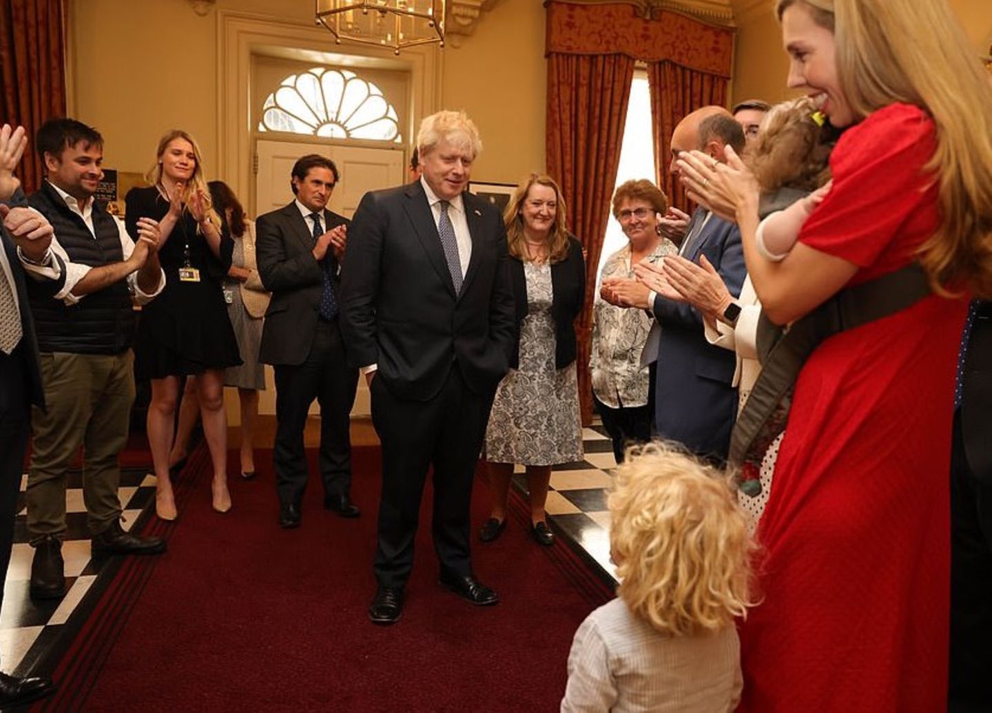 Boris Johnson steht in der Mitte eines Raumes, um ihn herum viele andere Männer und Frauen, die applaudieren