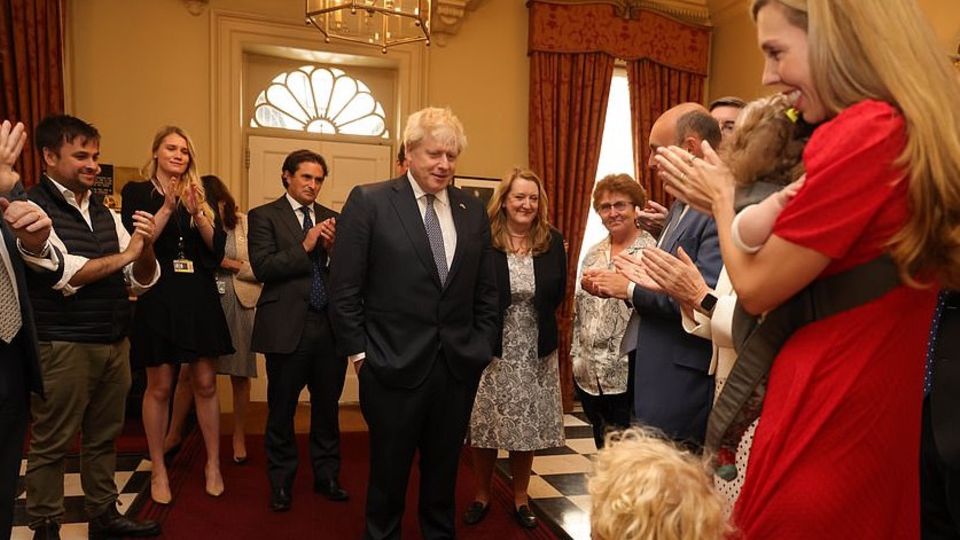 Boris Johnson steht in der Mitte eines Raumes, um ihn herum viele andere Männer und Frauen, die applaudieren