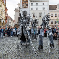 Drei Stelzenartisten laufen in Silber gekleidet über einen Marktplatz