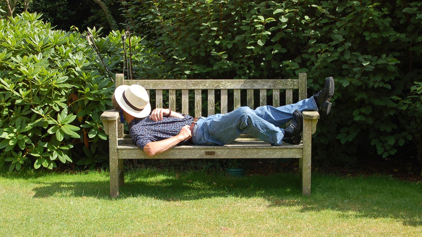 Ein Mann ruht sich auf einer Bank aus. Sein Gesicht schützt er mit einem Hut vor der Sonne.
