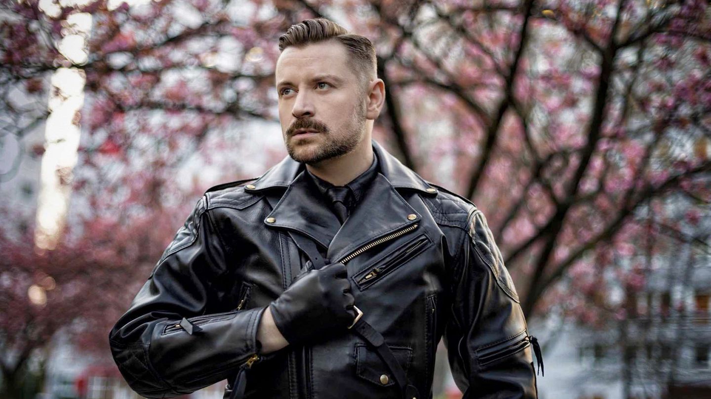 Ein Mann in Lederjacke und mit einem Lederhandschuh steht vor einem Baum