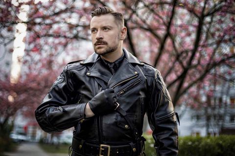Ein Mann in Lederjacke und mit einem Lederhandschuh steht vor einem Baum