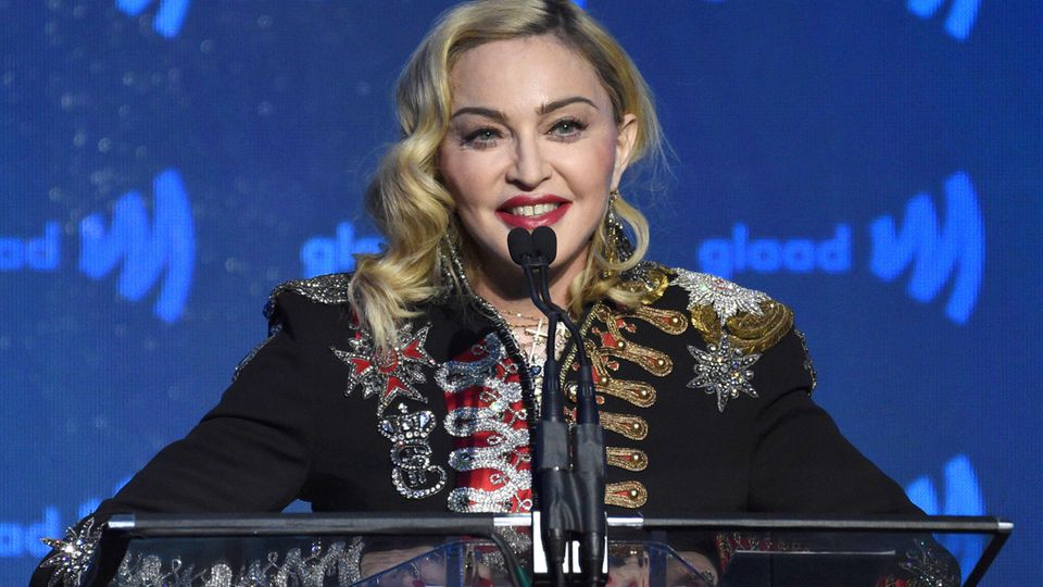 Madonna steht auf einer Bühne mit blauem Hintergrund und spricht in drei Mikrofone