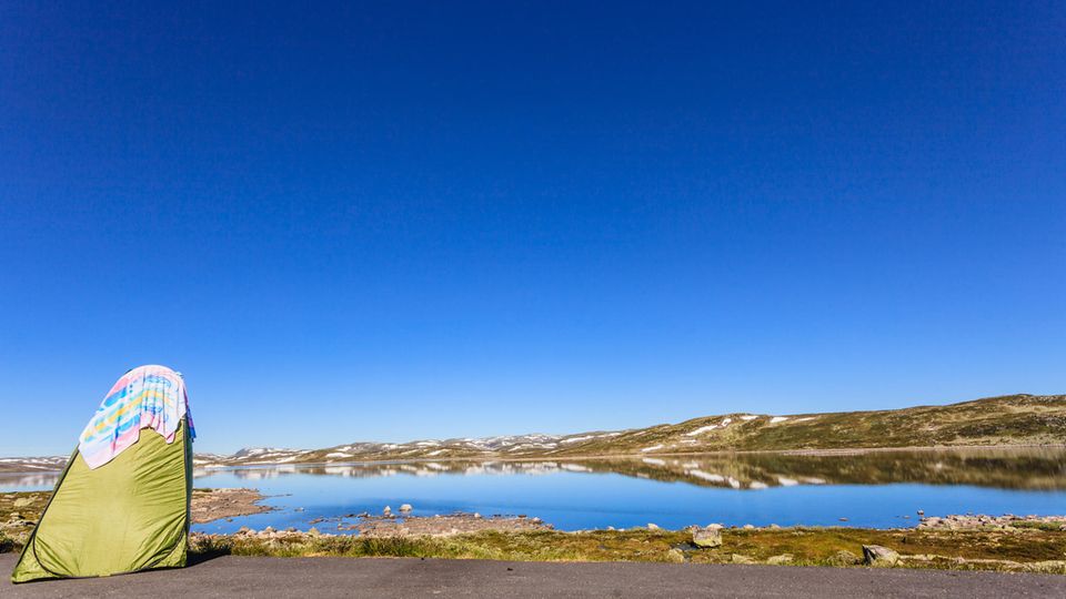 Ein Duschzelt steht an einem See. Im Hintergrund sind Hügel