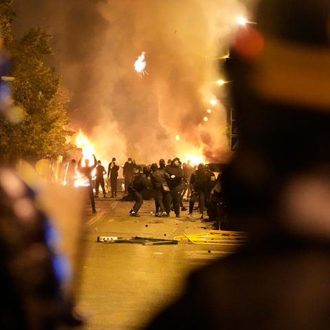 Polizisten mit Schutzschildern stehen Jugendlichen gegenüber. Dahinter brennt es.