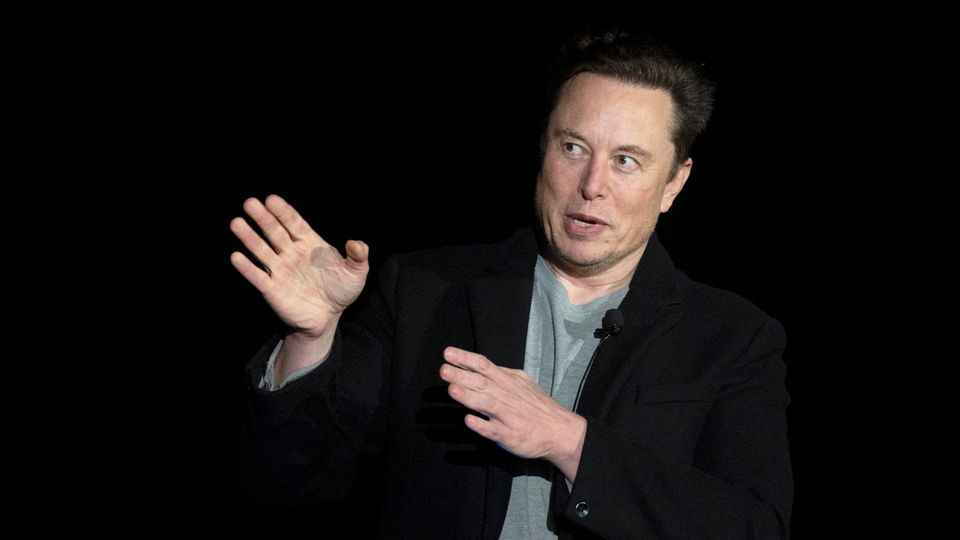 Elon Musk zeigt mit beiden Händen nach rechts und schaut mit großen Augen in die Richtung