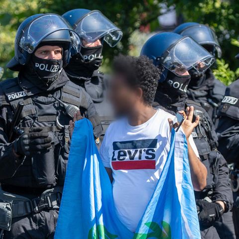 Ein Mann mit einer Flagge in den erhobenen Händen wendet sich einer Polizei-Kette zu