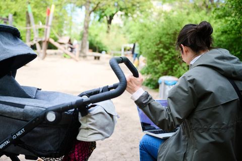 Eine Mutter am Laptop, während sie neben dem Kinderwagen auf dem Spielplatz sitzt