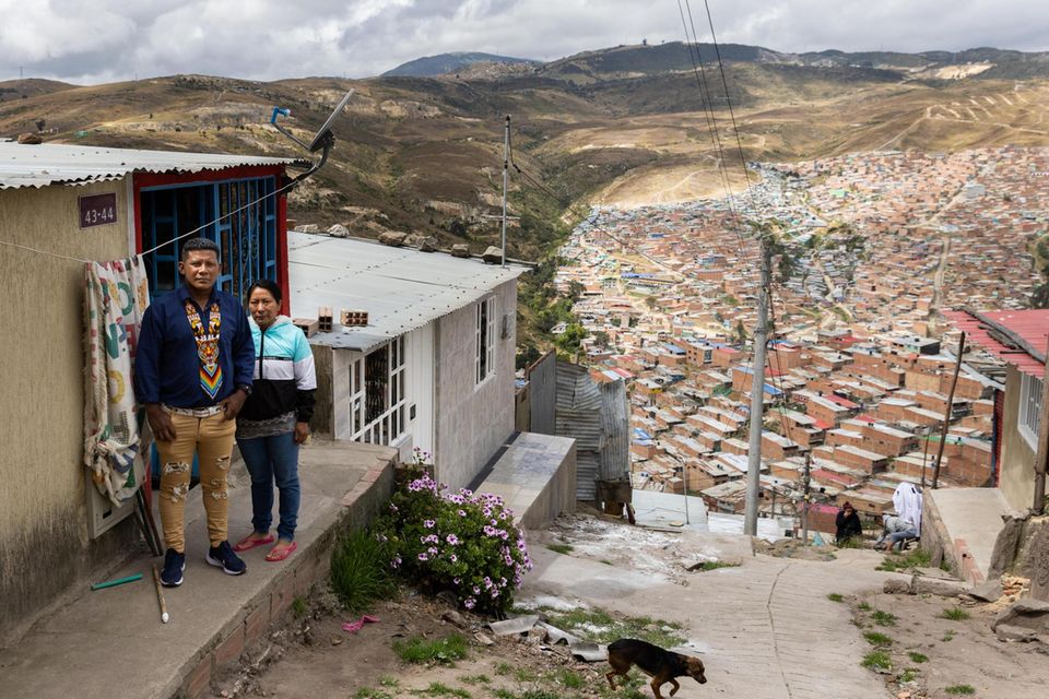 Zwei Menschen vor einer kleinen Hütte, im Hintergrund eine Siedlung