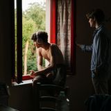 Ein junger Mann sitzt mit Kopfhörern am Fenster, daneben steht ein anderer Mann