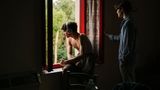 Ein junger Mann sitzt mit Kopfhörern am Fenster, daneben steht ein anderer Mann