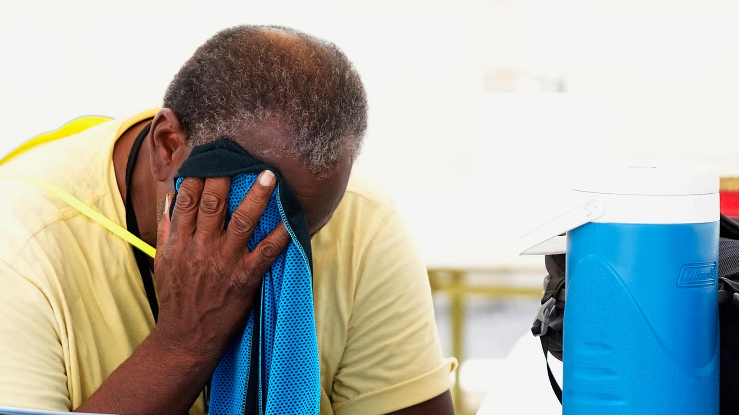 Ein Mann im gelben T-Shirt sitzt in der Sommerhitze und wischt sich mit einem blauen Handtuch Schweiß aus dem Gesicht