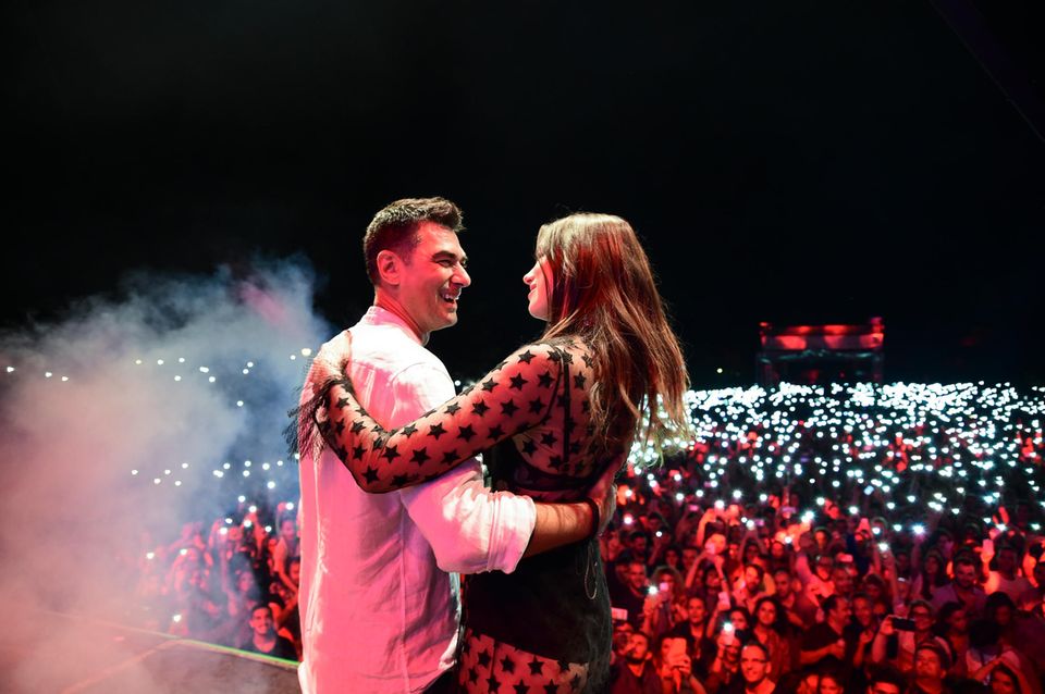 Dua Lipa and her father hug themselves on a stage in front of an audience