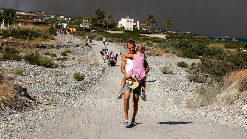 Ein Mann trägt auf der Ulraubsinsel Rhodos ein Kind, als sie ein Waldbrandgebiet verlassen