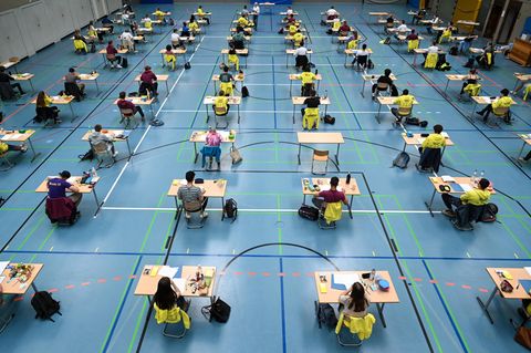 Mehrere Abiturienten sitzen bei einer Prüfung mit Abstand zueinander an Tischen in Turnhallen