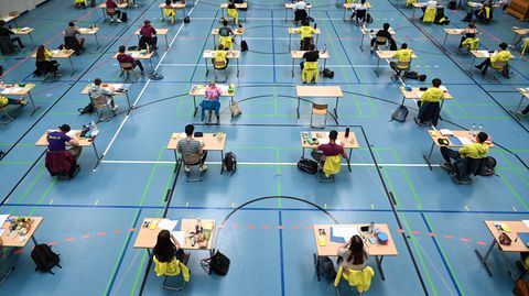 Mehrere Abiturienten sitzen bei einer Prüfung mit Abstand zueinander an Tischen in Turnhallen
