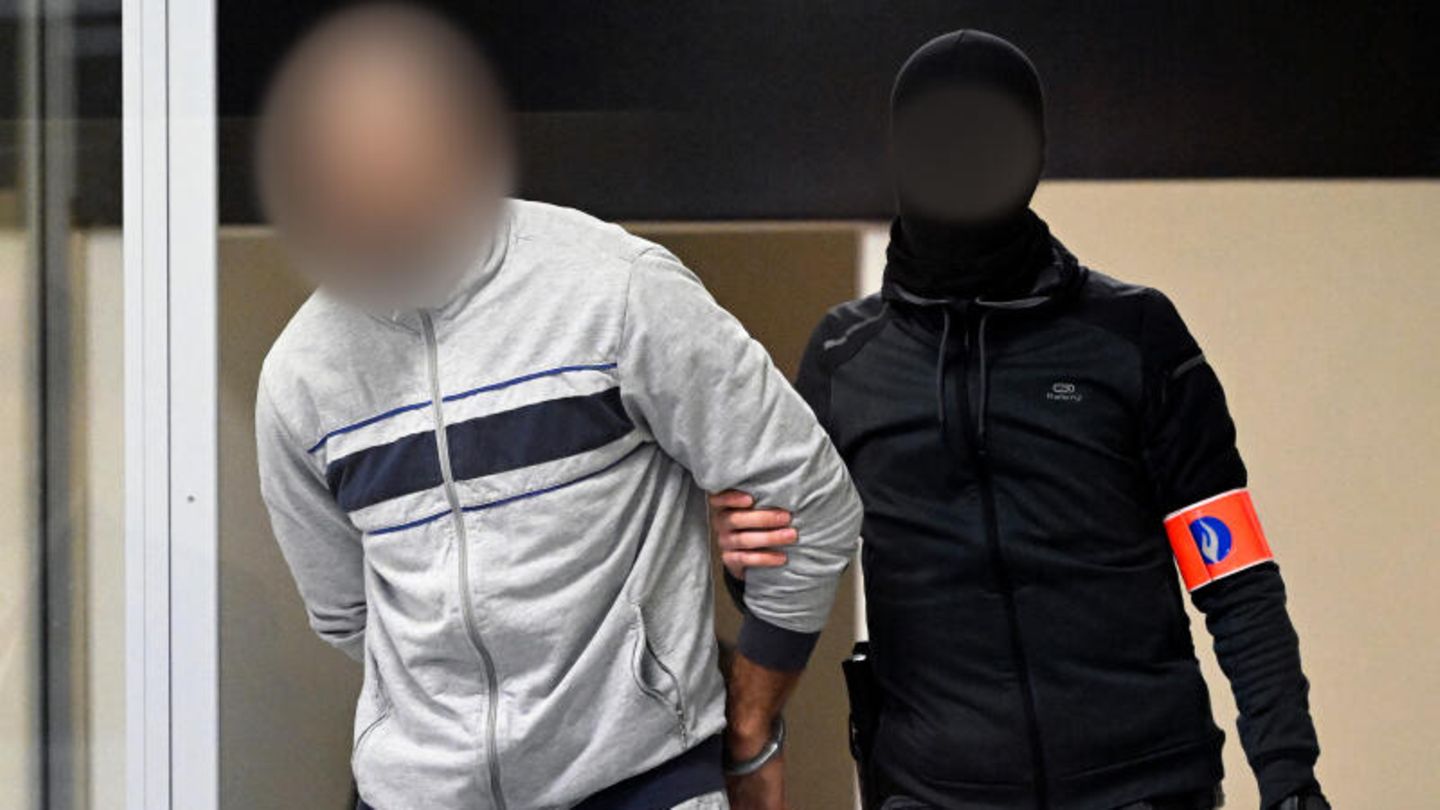 Ein vermummter Spezialpolizist bringt einen der Angeklagten in den Glaskasten in den Verhandlungssaal (Archivbild)