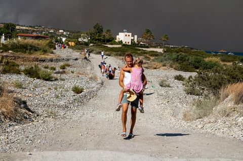 Ein Mann trägt auf Rhodos ein Kind eine Schotterstraße herunter, während schwarzer Rauch eines Waldbrandes aufsteigt