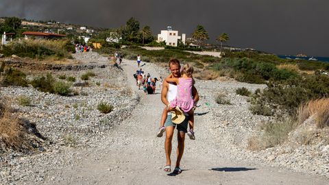 Ein Mann trägt auf Rhodos ein Kind eine Schotterstraße herunter, während schwarzer Rauch eines Waldbrandes aufsteigt