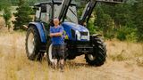 Mann mit blauem T-Shirt und dunkelgrauer Hose steht vor blauem Traktor
