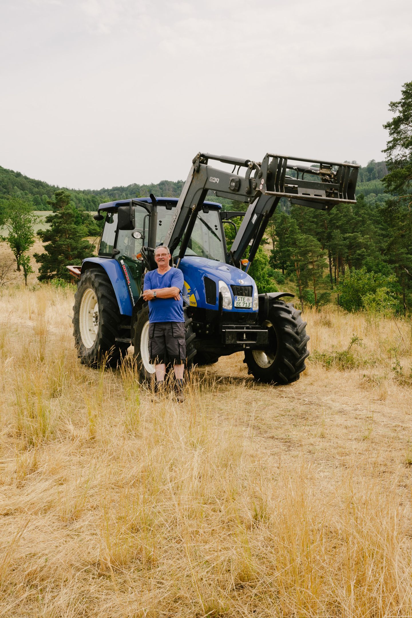 Mann mit blauem T-Shirt und dunkelgrauer Hose steht vor blauem Traktor