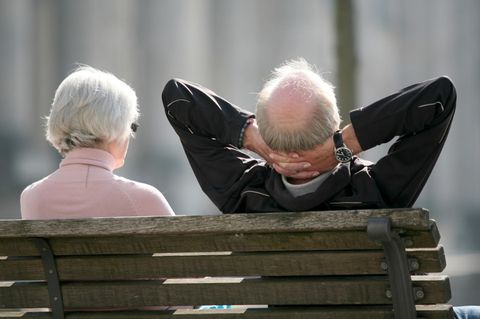 Von hinten sind eine weißhaarige Frau und ein weißhaariger Mann mit Halbglatze zu sehen, die auf einer Bank in der Sonne sitzen