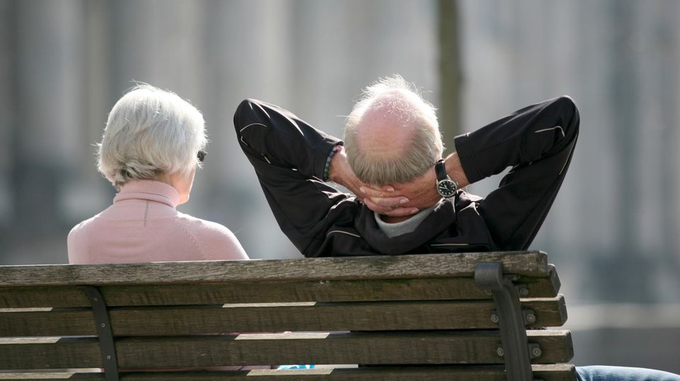 Von hinten sind eine weißhaarige Frau und ein weißhaariger Mann mit Halbglatze zu sehen, die auf einer Bank in der Sonne sitzen