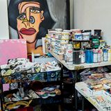 Auf einem Regal in der Ecke stapeln sich Farbtöpfe, Schachteln mit Öl-Malstiften und selbstgemachte Skulpturen. An der Wand lehnt ein Gemälde, das ein Porträt einer Frau mit Nasenring zeigt