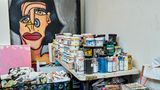 Auf einem Regal in der Ecke stapeln sich Farbtöpfe, Schachteln mit Öl-Malstiften und selbstgemachte Skulpturen. An der Wand lehnt ein Gemälde, das ein Porträt einer Frau mit Nasenring zeigt