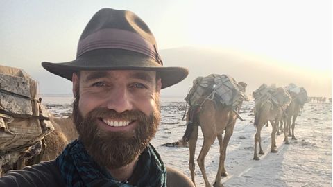 Ein Mann mit Hut und dunklem Vollbart guckt in die Kamera und lächelt, hinter ihm stehen bepackte Kamele