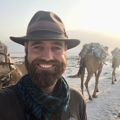 Ein Mann mit Hut und dunklem Vollbart guckt in die Kamera und lächelt, hinter ihm stehen bepackte Kamele