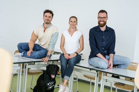 Drei junge Lehrer sitzen auf einem Pult, zu Füßen der Schulhund, ein schwarzer Labrador