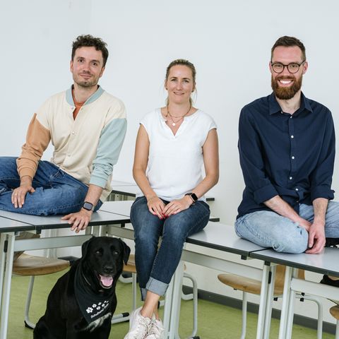 Drei junge Lehrer sitzen auf einem Pult, zu Füßen der Schulhund, ein schwarzer Labrador