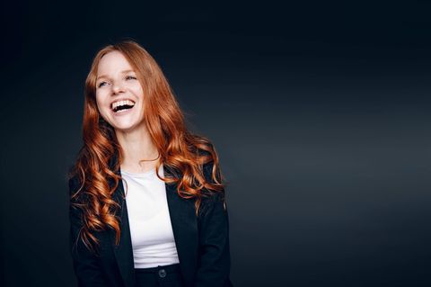 Eine rothaarige Frau mit großem Lachen vor einem dunklen Hintergrund