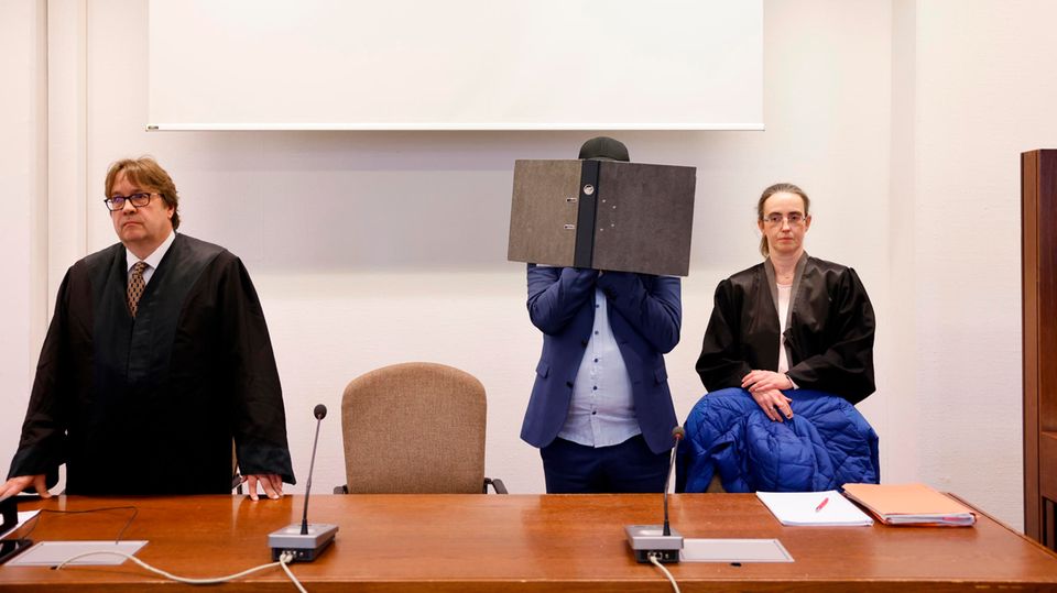 Der Angeklagte steht zwischen seinen Rechtsanwälten, während er sein Gesicht hinter einem Aktenordner verbirgt