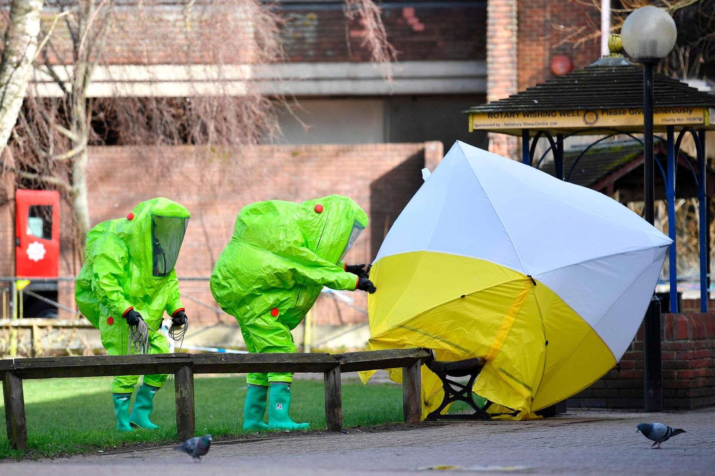 Zwei Menschen in grünen Schutzanzügen und ein Sichtschutz vor der Parkbank Zwei Menschen in grünen Schutzanzügen und ein Sichtschutz vor der Parkbank
