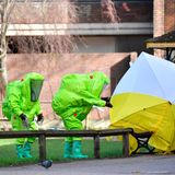 Zwei Menschen in grünen Schutzanzügen und ein Sichtschutz vor der Parkbank