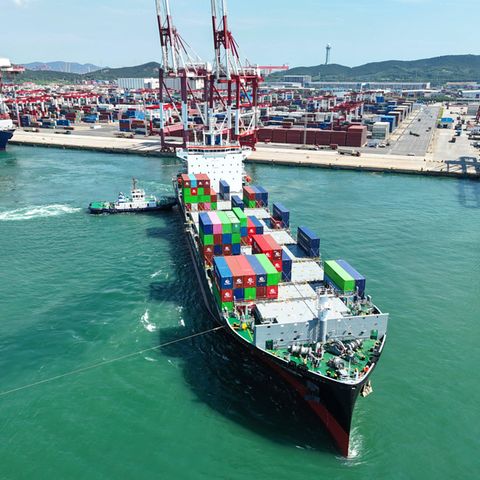 Ein Containerschiff verlässt den Hafen von Quingdao in China, im Hintergrund Kaianlagen und Hafenkräne