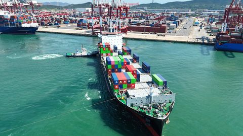 Ein Containerschiff verlässt den Hafen von Quingdao in China, im Hintergrund Kaianlagen und Hafenkräne