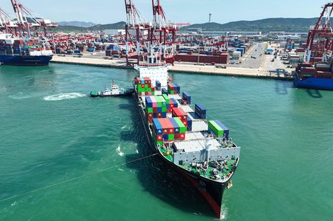 Ein Containerschiff verlässt den Hafen von Quingdao in China, im Hintergrund Kaianlagen und Hafenkräne